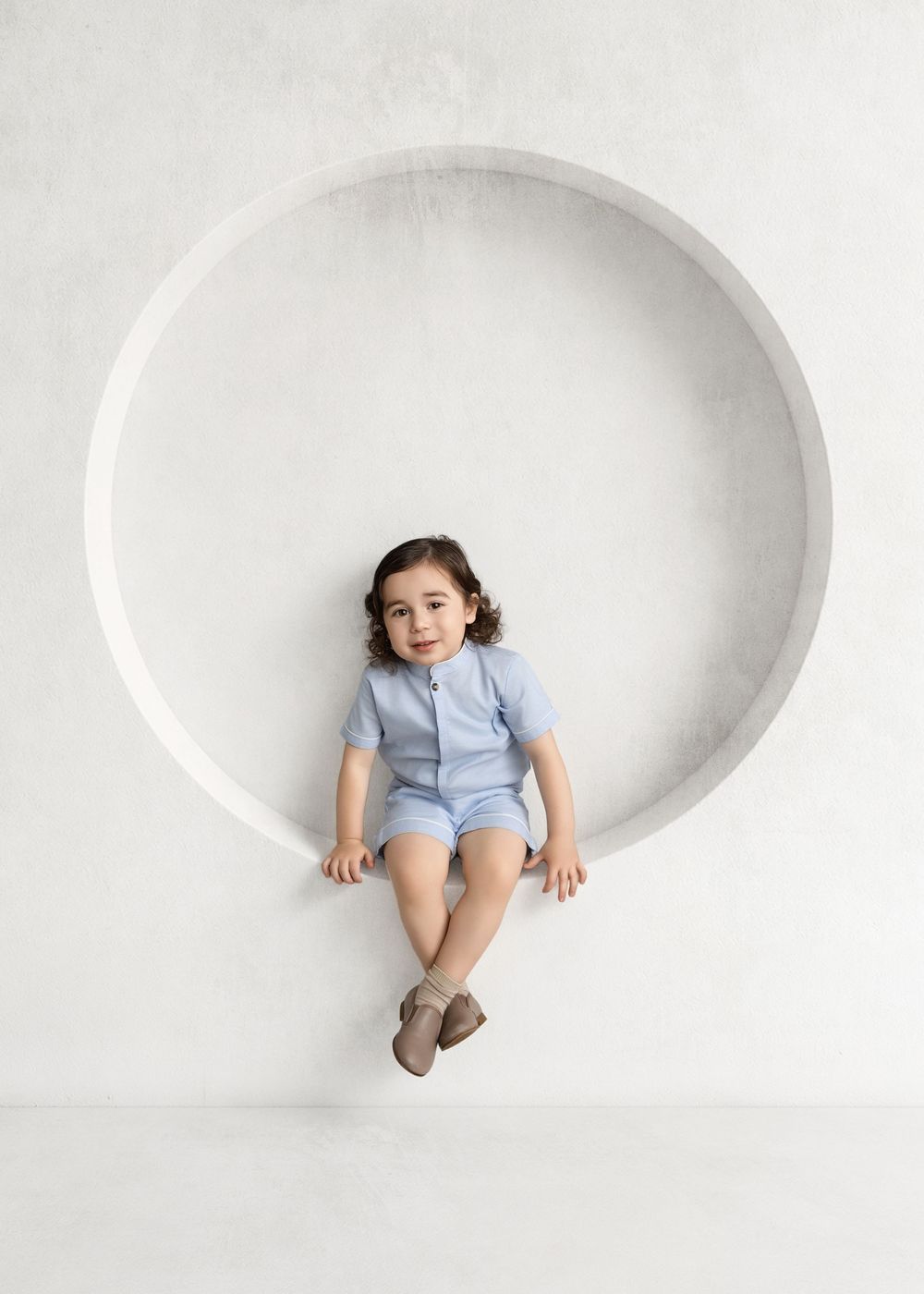 Young child in blue shorts set sitting in a circular wall niche, smiling at camera against white background.