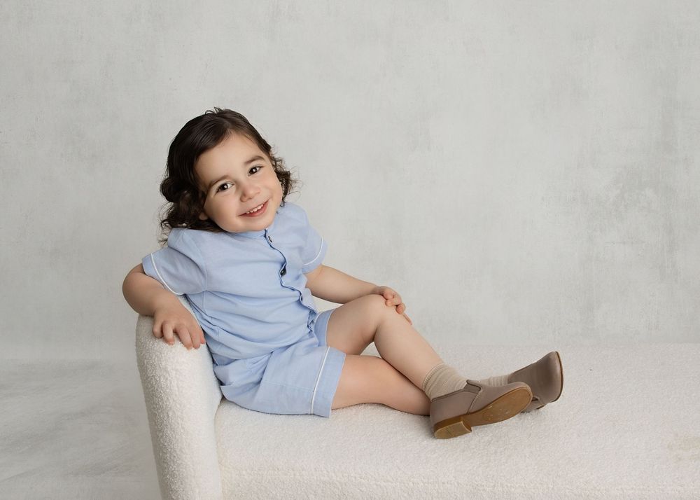 Toddler girl in light blue romper sitting on white chair, smiling, wearing tan shoes, against gray background.
