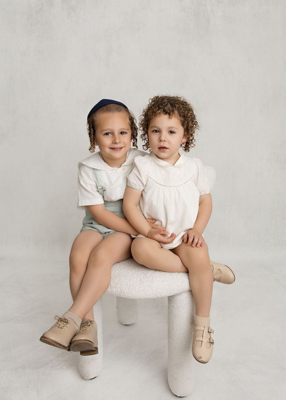 Two young children in white outfits sit together on a small stool, smiling for a professional portrait photo.