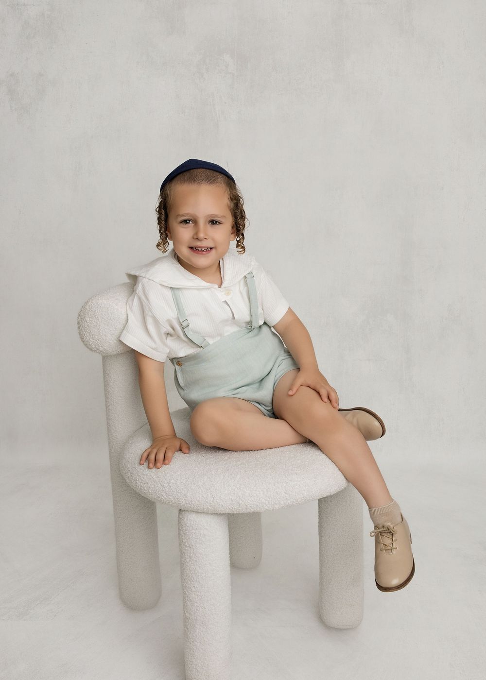 Young child sitting on a white boucle chair, wearing mint overalls and white top, smiling for a portrait photo.