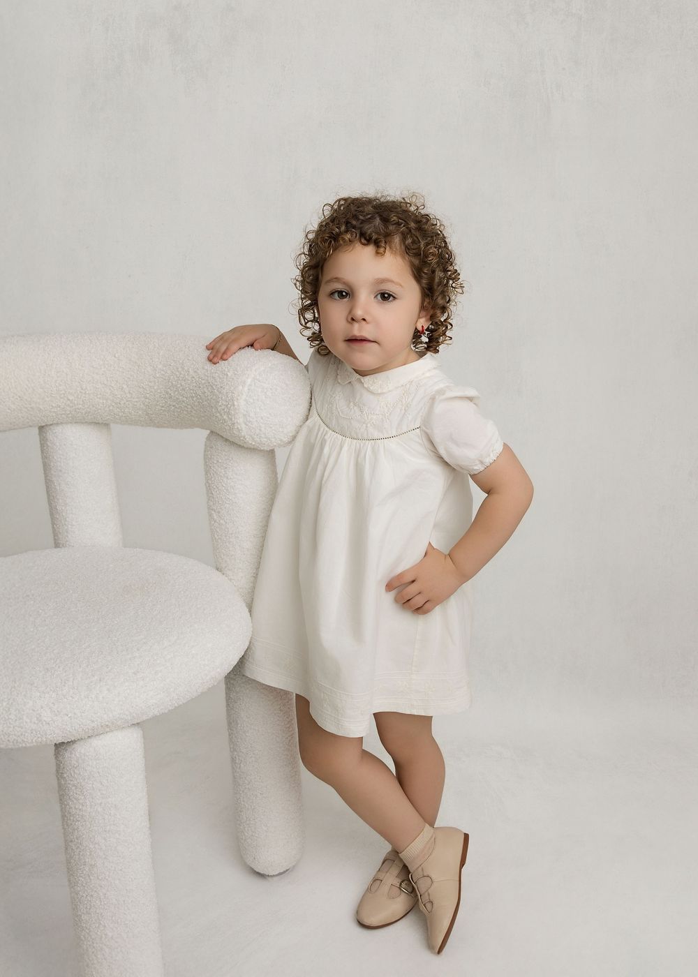 Toddler girl in white dress with puff sleeves and beige shoes poses beside a white boucle chair.