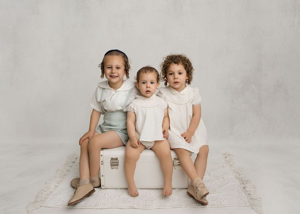 Three young children in white outfits sitting together on a white trunk against a light gray background.