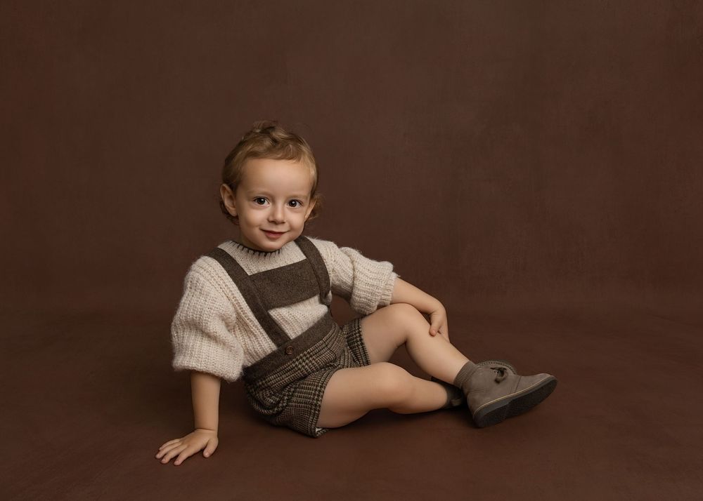 Adorable toddler boy sitting in brown overalls and knit sweater, smiling at camera against warm brown backdrop.