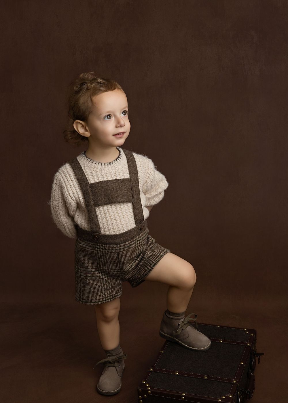 Young child in vintage-style knit sweater and plaid suspender shorts poses on antique suitcase against brown backdrop.