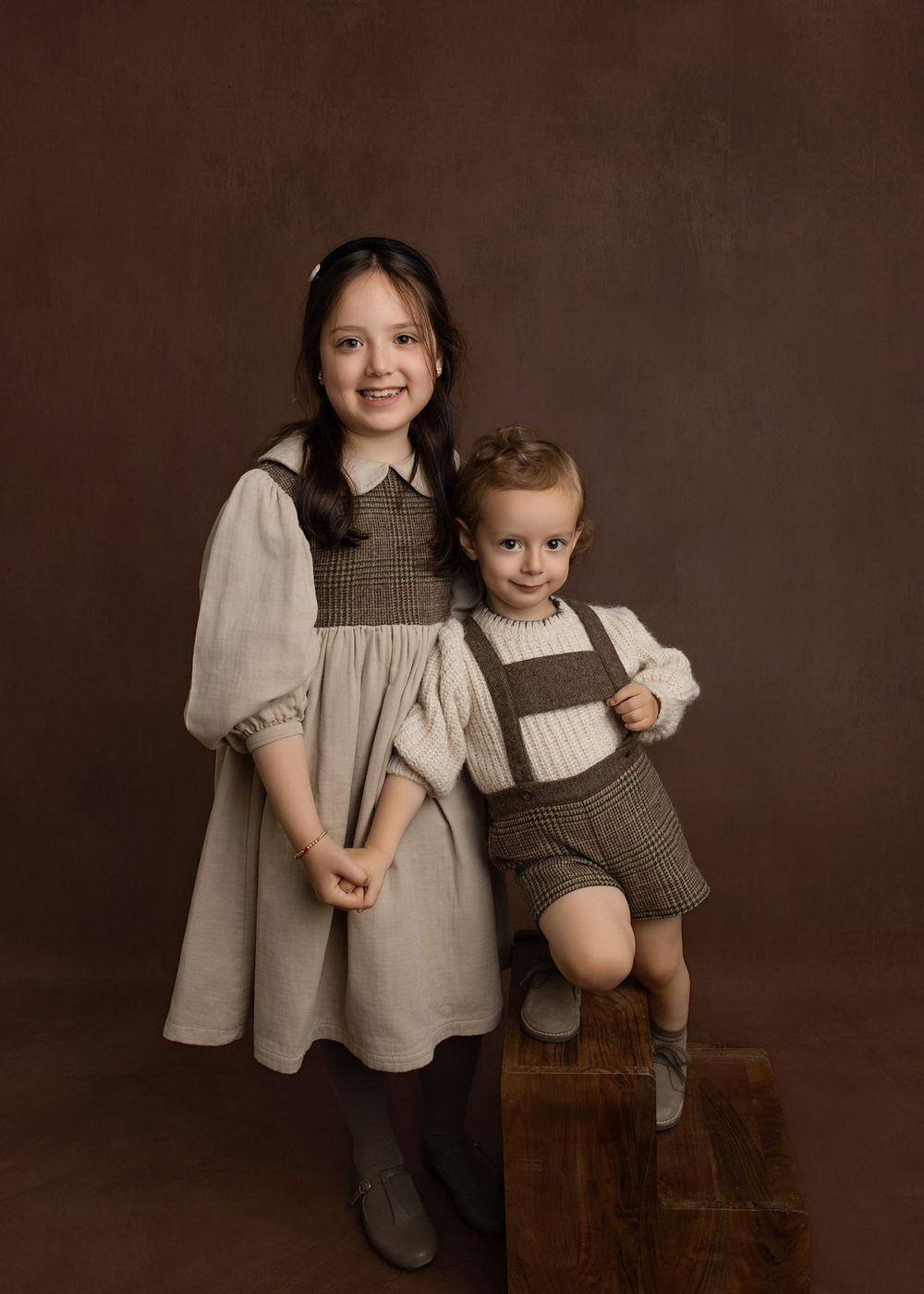 Two young siblings in vintage-style coordinating outfits pose together for a classic studio portrait against a brown background.