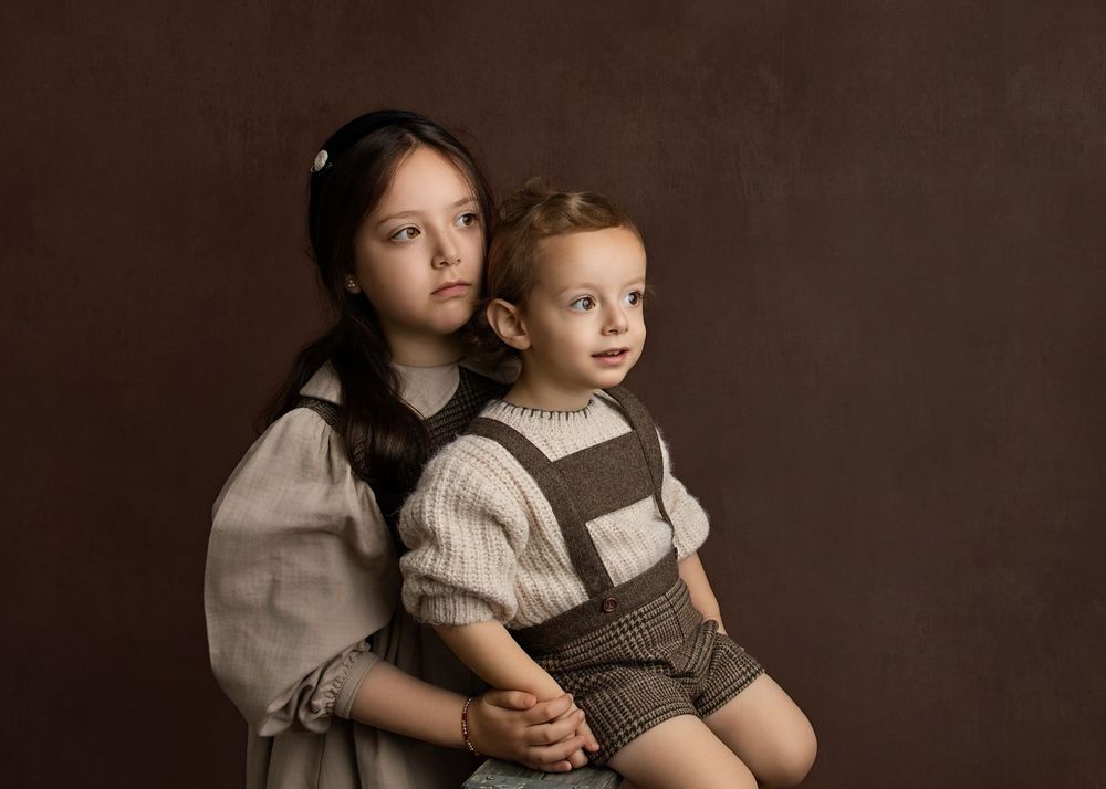Two children pose for a vintage-style portrait, an older girl hugging a younger boy against a brown backdrop.