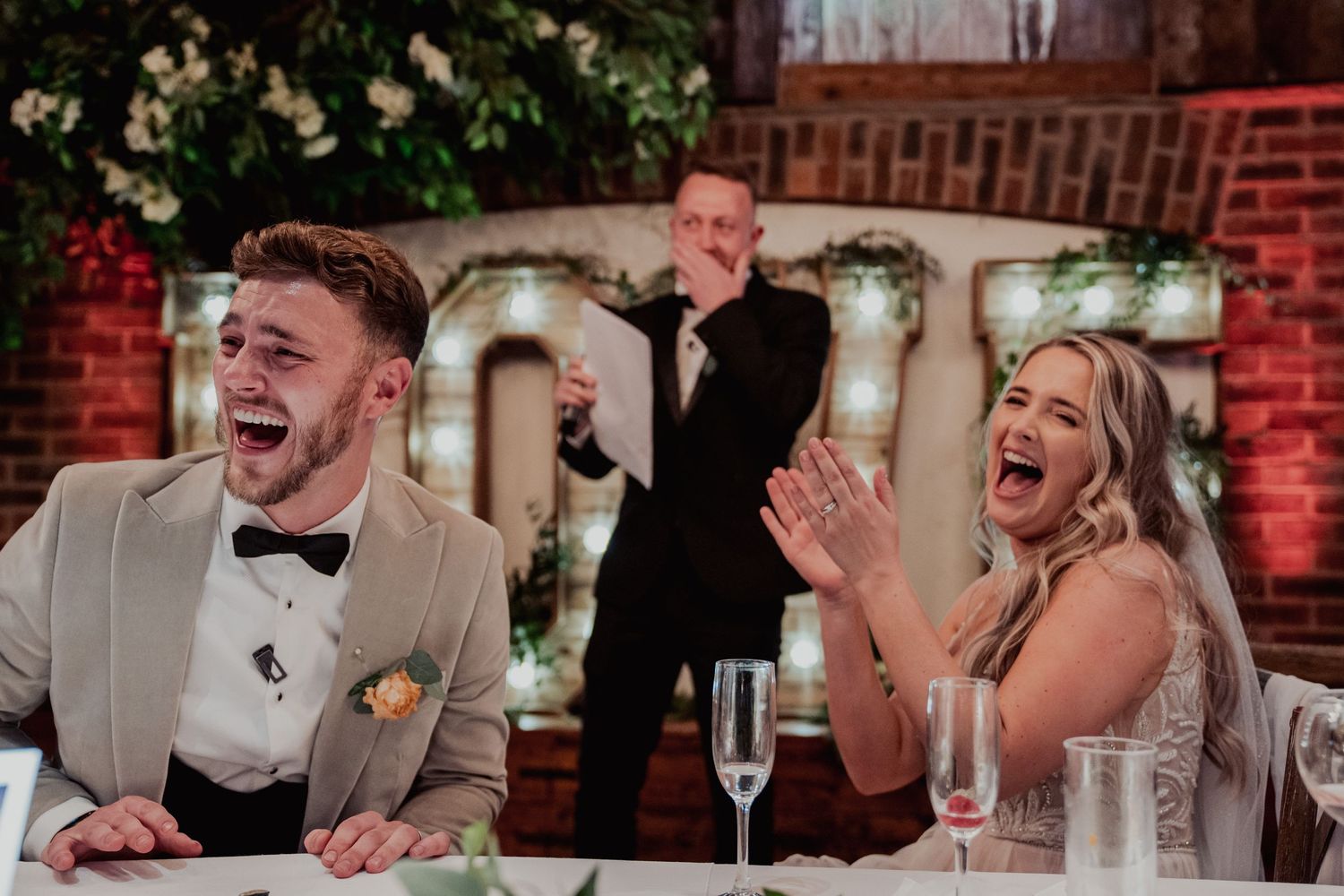 Wedding party guests laugh joyfully during speeches at reception with white floral decor and red lighting.