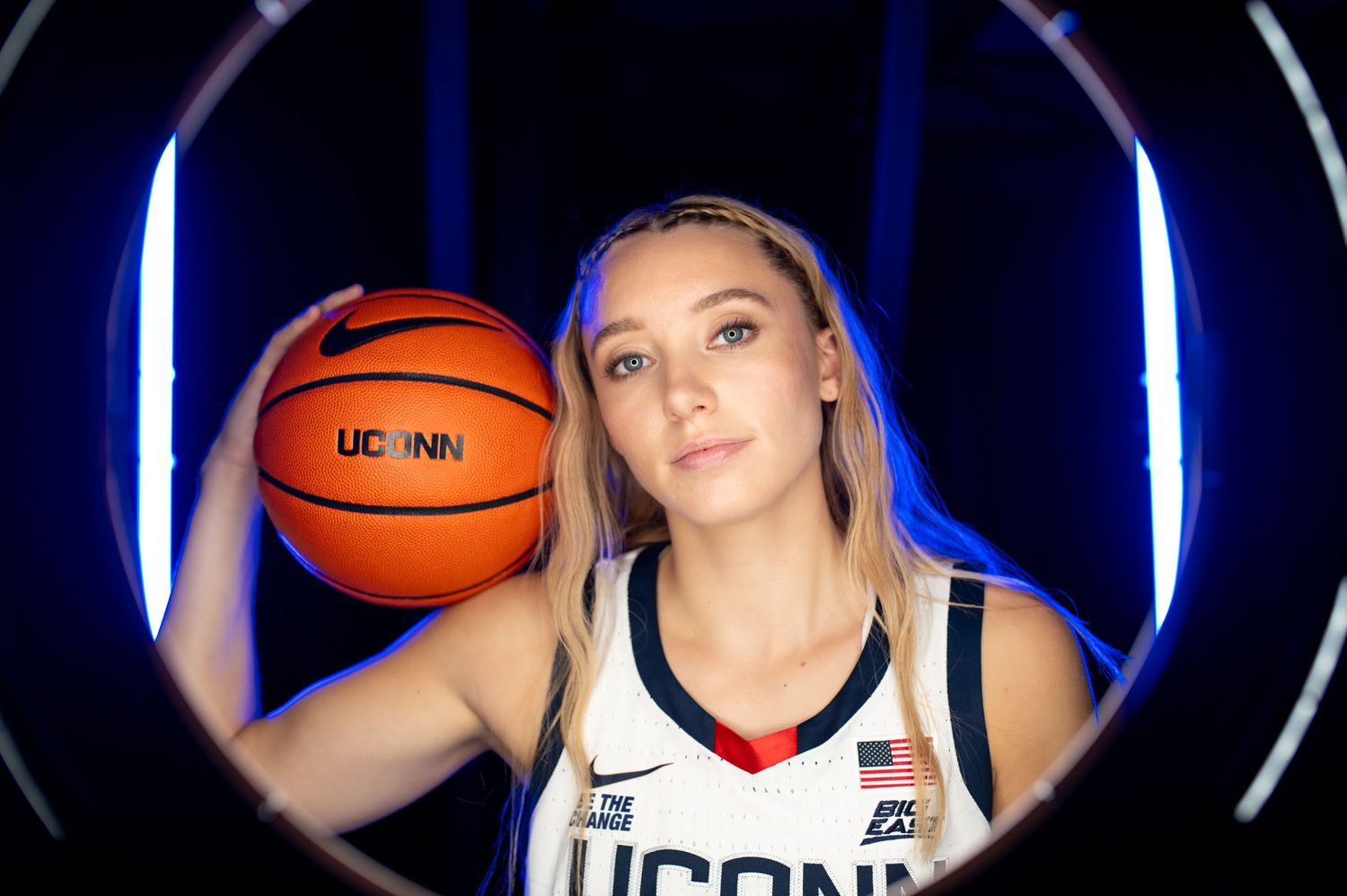 Basketball player in UCONN uniform posing with basketball against dark background with blue lighting.