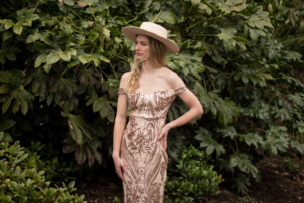 A figure in a champagne sequined gown and wide-brim hat poses against lush green foliage.