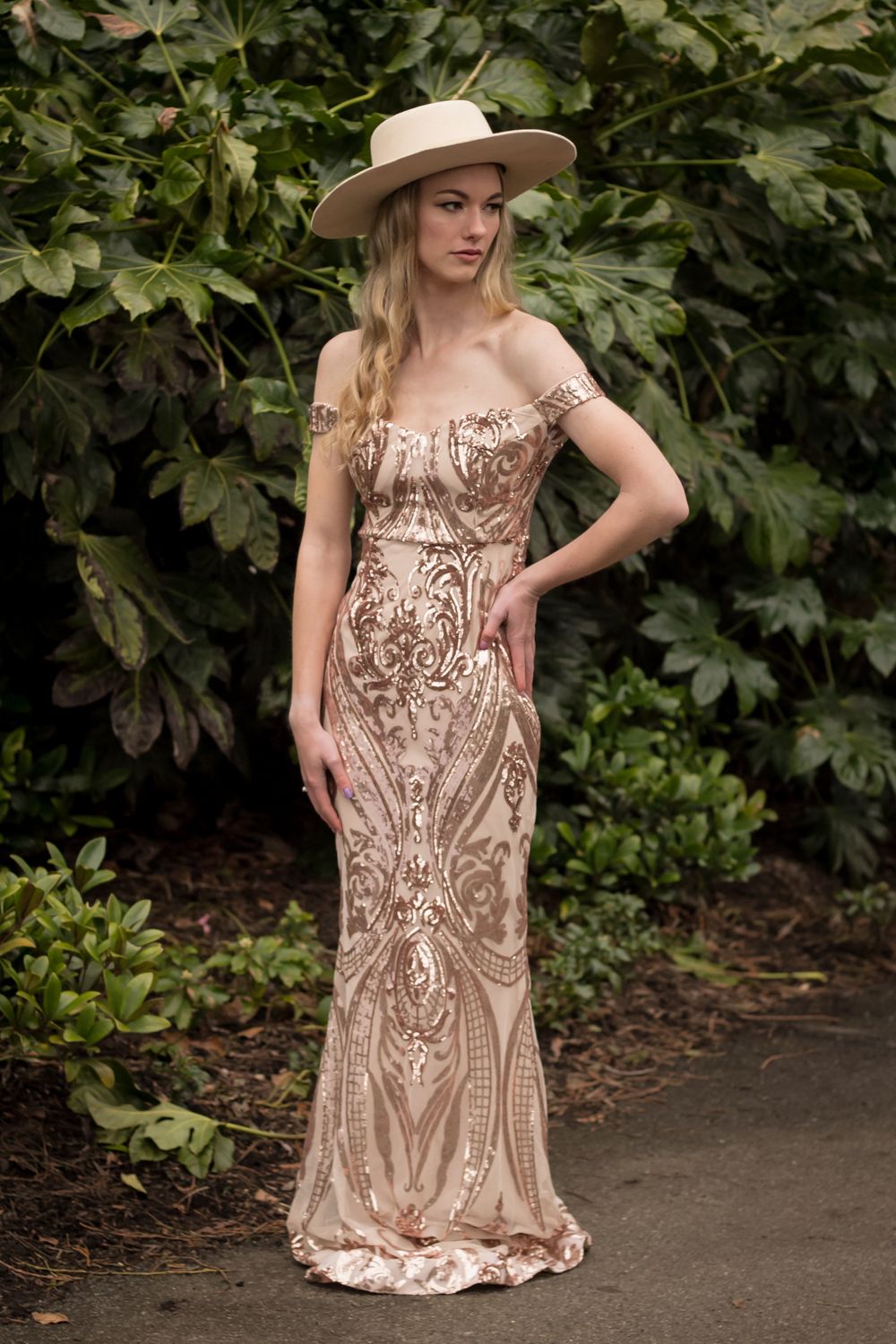 Elegant full-length portrait in beaded champagne gown and wide-brimmed hat against garden foliage.