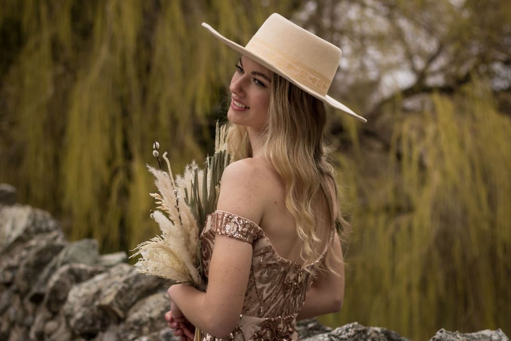 Romantic portrait of person in vintage dress holding dried flowers against willow tree backdrop.