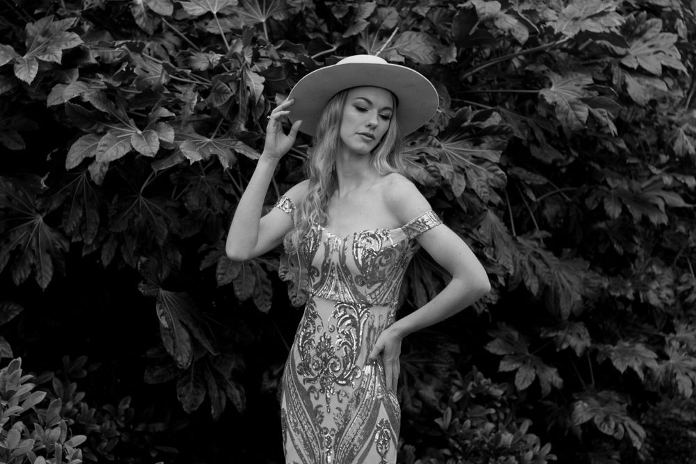 Black and white portrait in ornate gown and hat with natural foliage background.