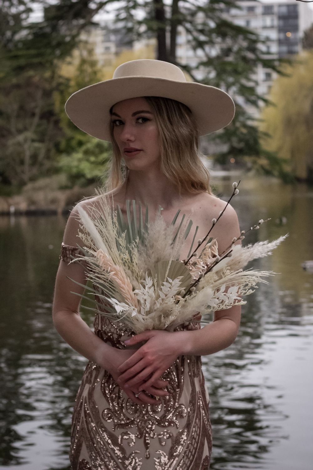 Figure in beige hat holds dried flower bouquet against pond backdrop.