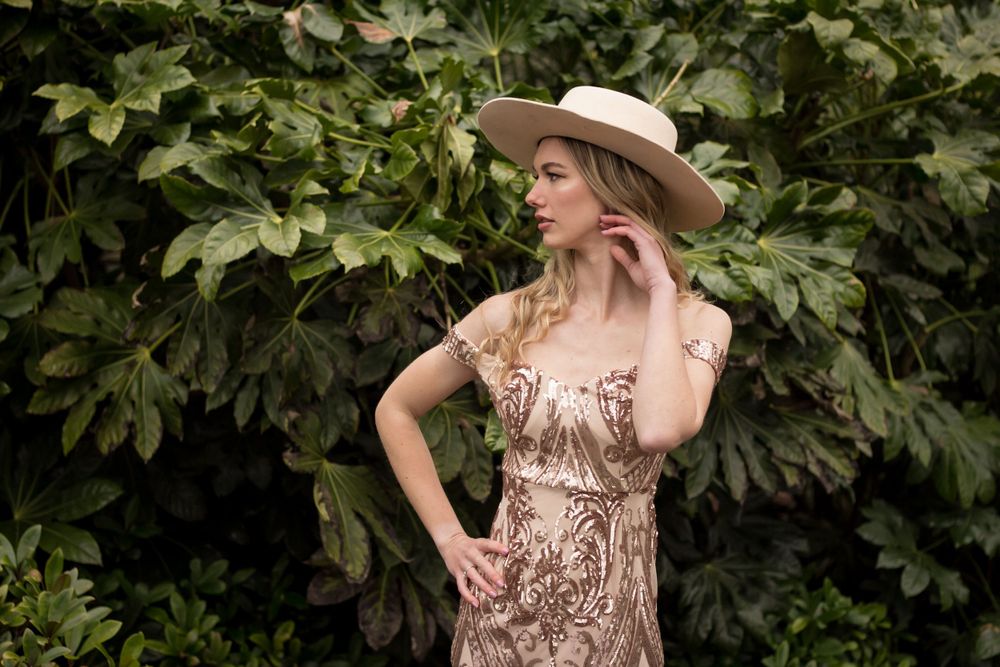 A graceful stance in a shimmering evening gown and neutral hat against garden greenery.