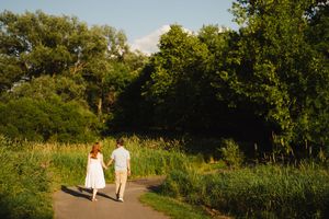 Two figures walking along a sunlit nature trail surrounded by lush greenery.