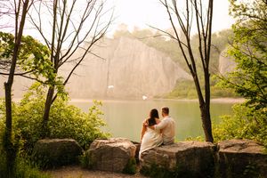 A couple shares a romantic moment on a stone wall surrounded by autumn trees at sunset.
