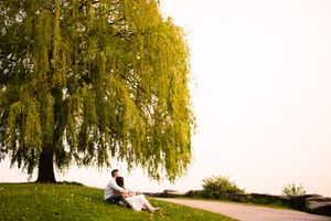 A couple sits beneath drooping willow trees near a tranquil lake.