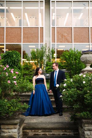 A couple poses in formal attire against a garden wall with blooming roses.