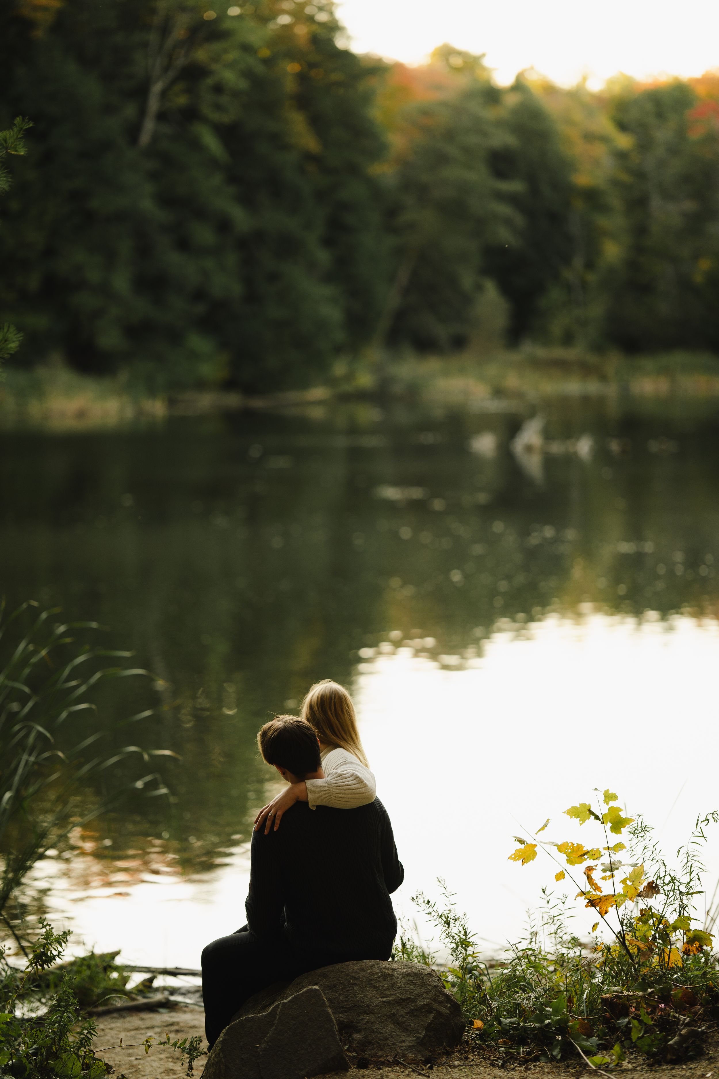 Couple sits by serene lake surrounded by autumn foliage at sunset.
