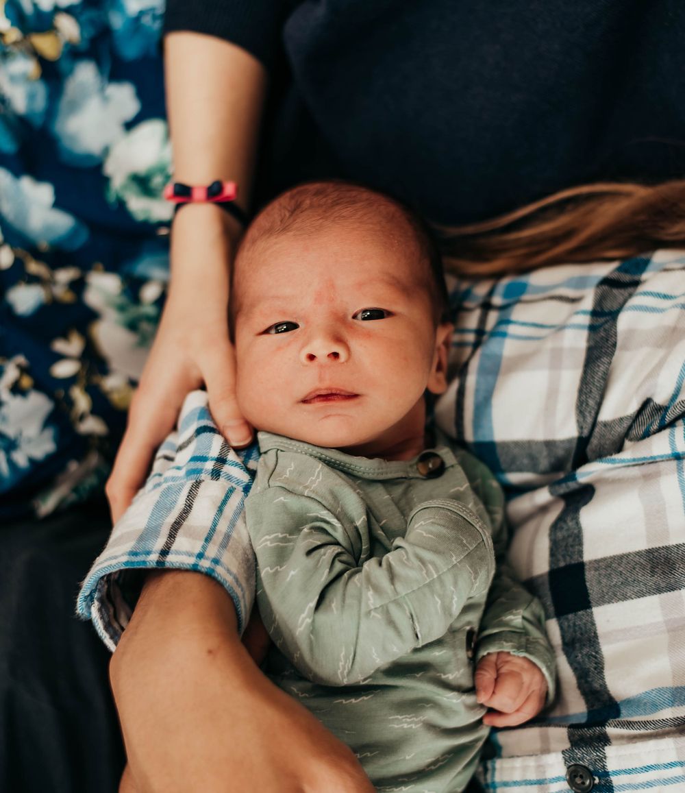 Alert newborn baby in green outfit resting on parent's lap in plaid clothing overhead view.