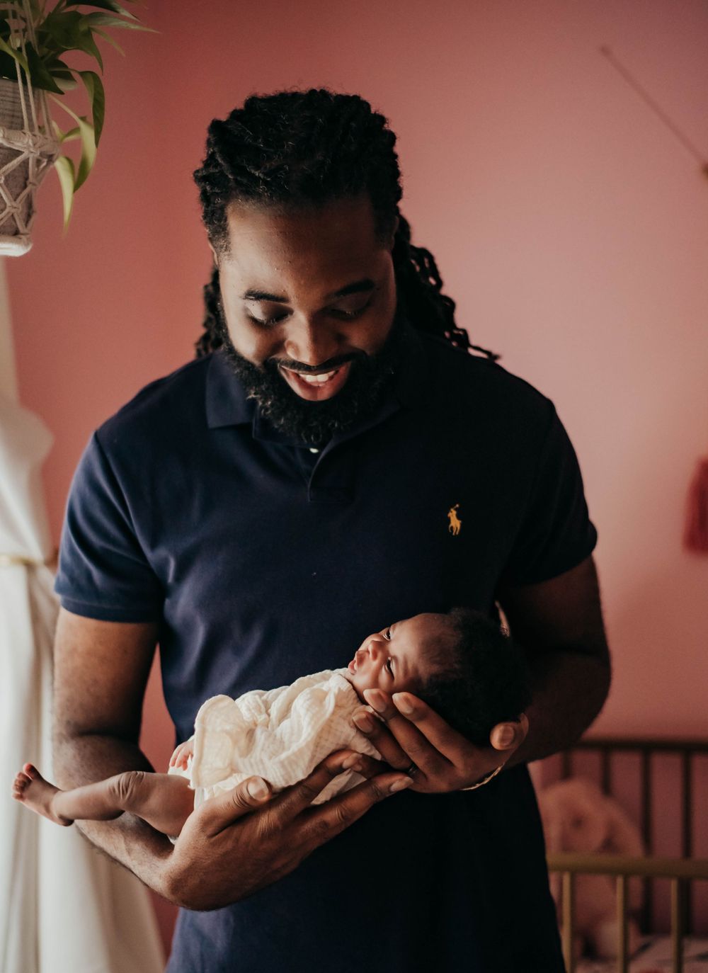 Smiling father with dreadlocks holding newborn baby in navy Ralph Lauren polo shirt in pink nursery.