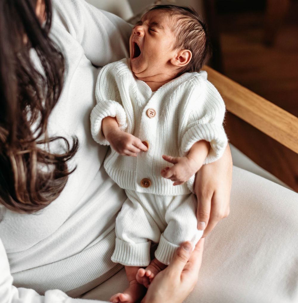 Newborn baby in white knit outfit yawning while being held by mother in bright natural light.