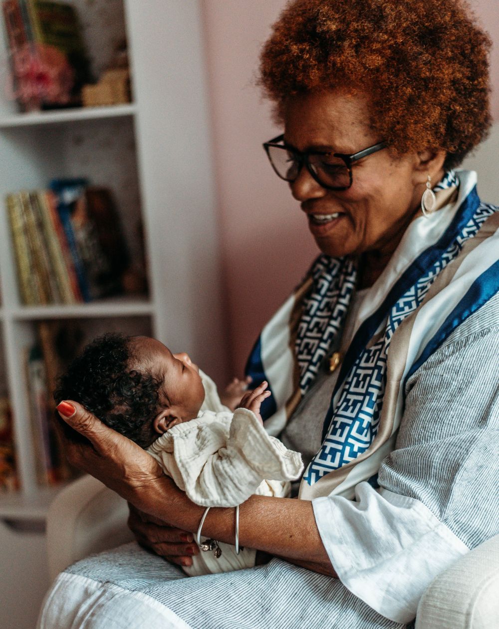 Grandmother with curly hair and glasses smiling warmly while holding newborn baby in cozy room.