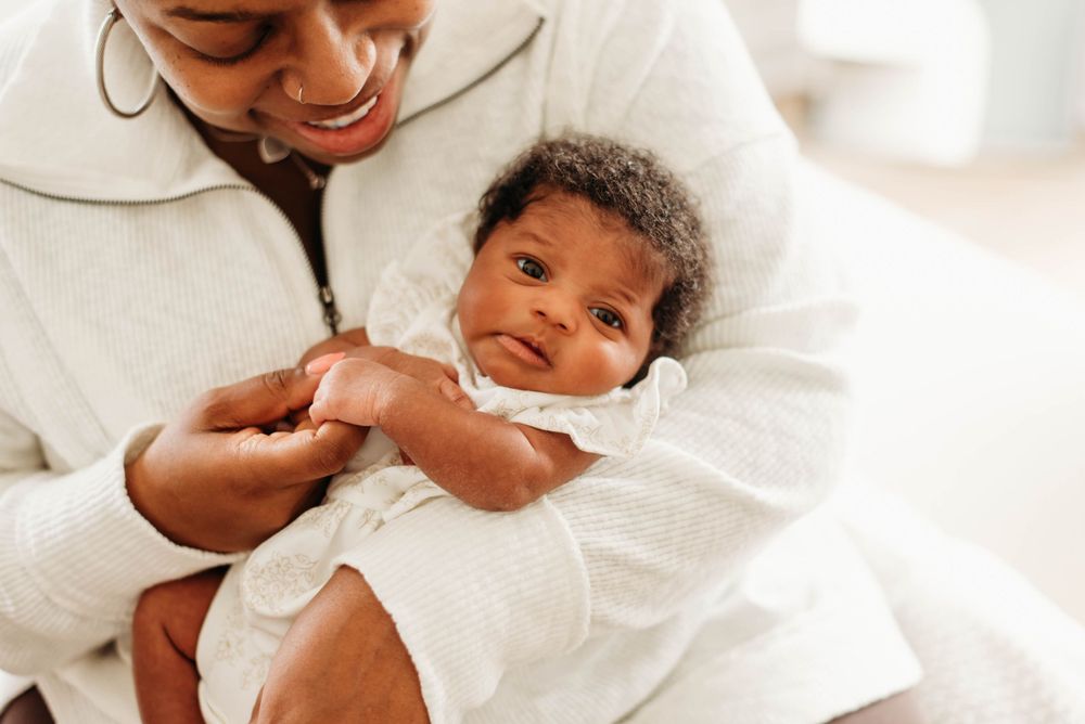 Happy mother in white outfit holding alert newborn baby with curly hair in soft natural light.