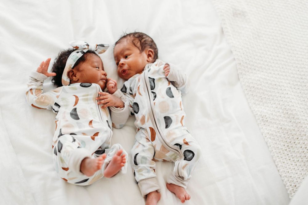 Twin newborn babies in matching patterned onesies sleeping side by side on white bedding.
