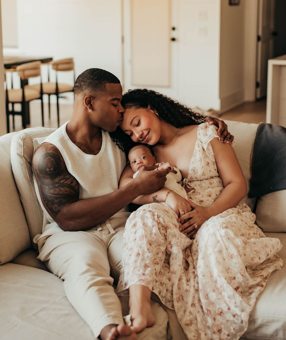 Young family with tattooed father kissing mothers head while holding newborn on cozy sofa.