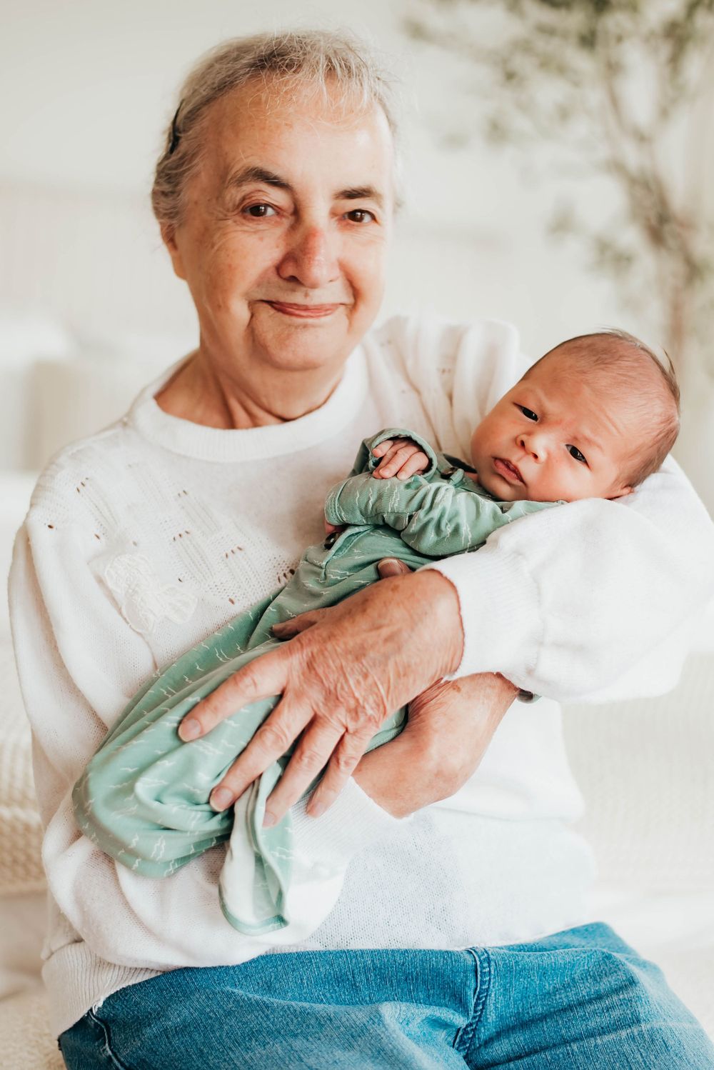 Smiling elderly grandfather in white sweater holding sleeping newborn wrapped in green blanket.
