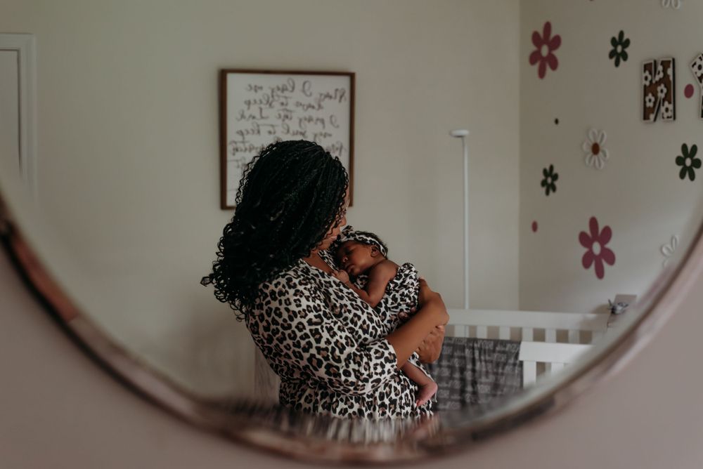 Mother holding newborn baby reflected in round mirror in floral nursery room.