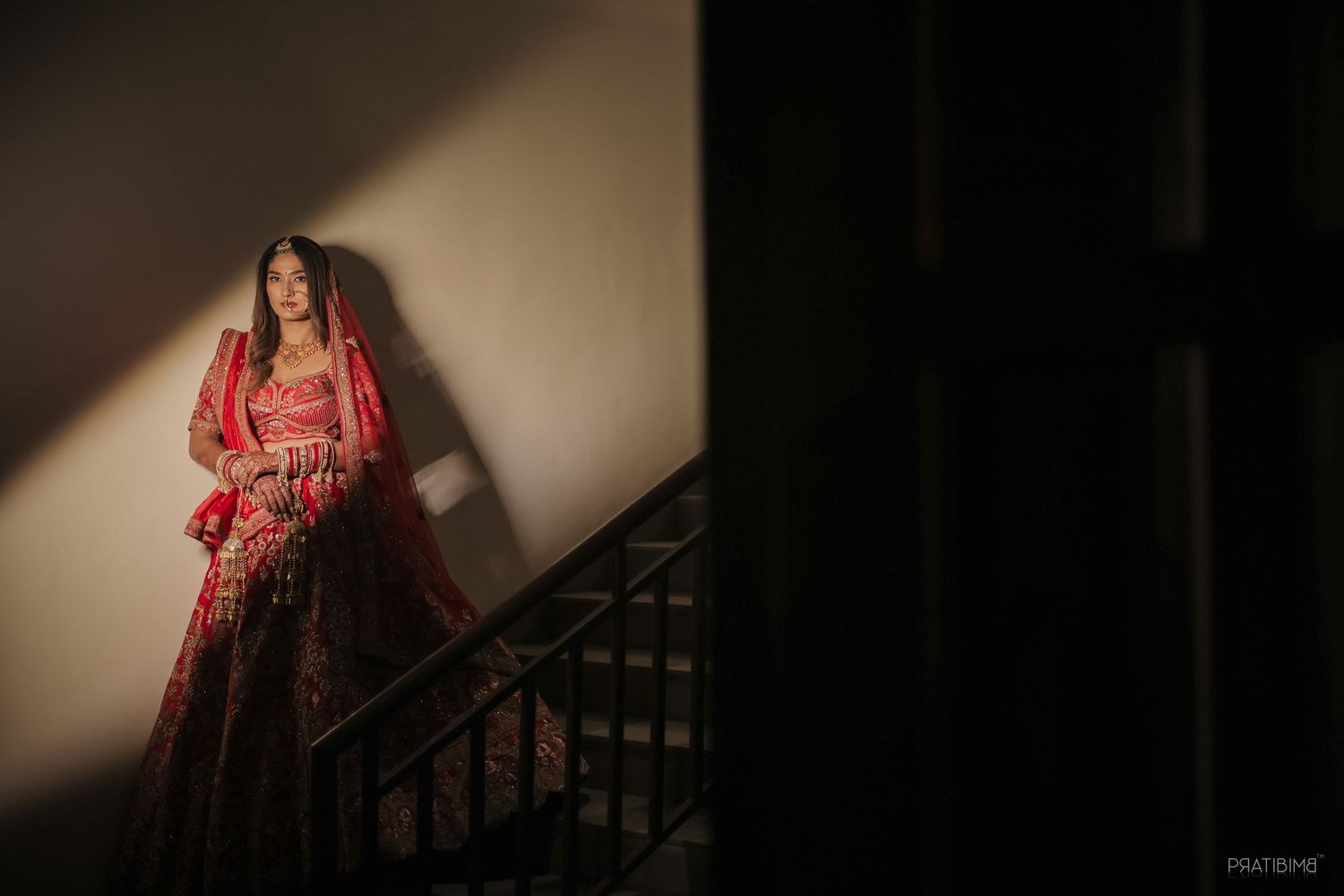 Bride in red bridal lehenga standing on staircase in dramatic light against white wall.