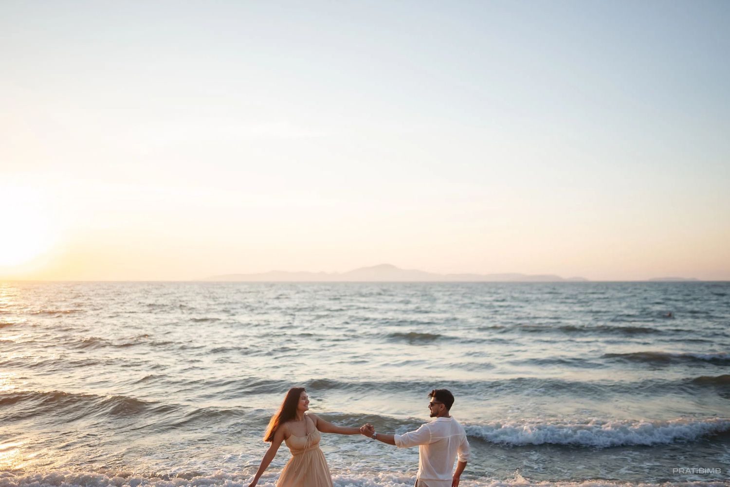 Couple holding hands on a sandy beach at sunset with calm ocean waves and distant mountains.