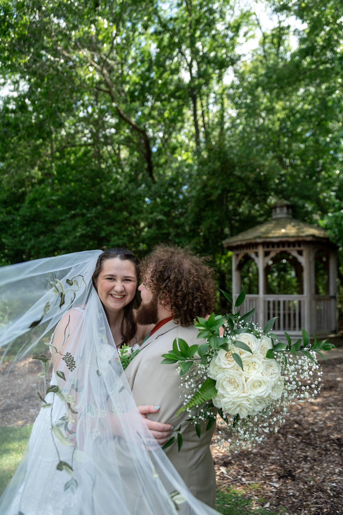 Bride smiling joyfully as groom kisses her cheek, holding white rose bouquet near a garden gazebo.