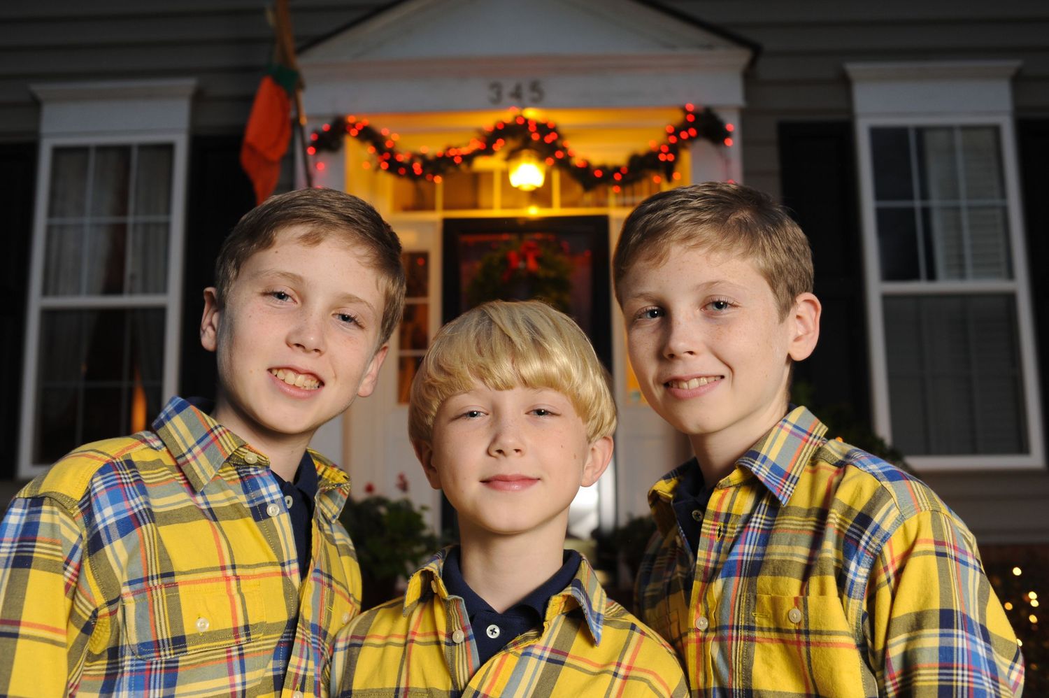 Three boys in matching yellow plaid shirts pose together outdoors in front of a holiday-decorated house at night.