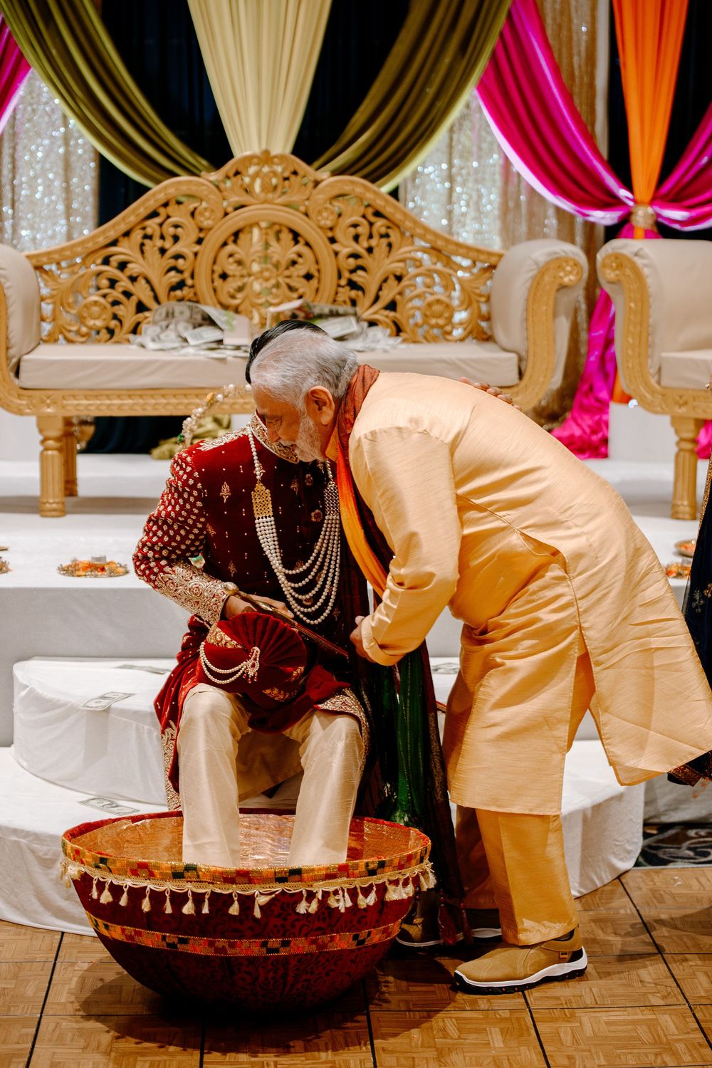 Traditional Indian wedding ceremony with groom in red sherwani having feet washed in decorative basin by elder in gold attire.