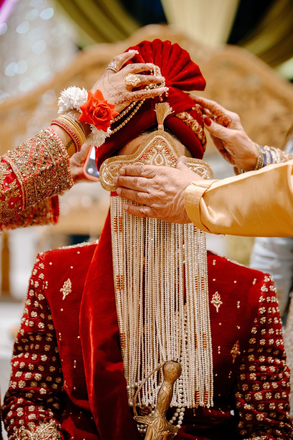 Groom in red sherwani and turban during Indian wedding ceremony with pearl sehra veil being placed.
