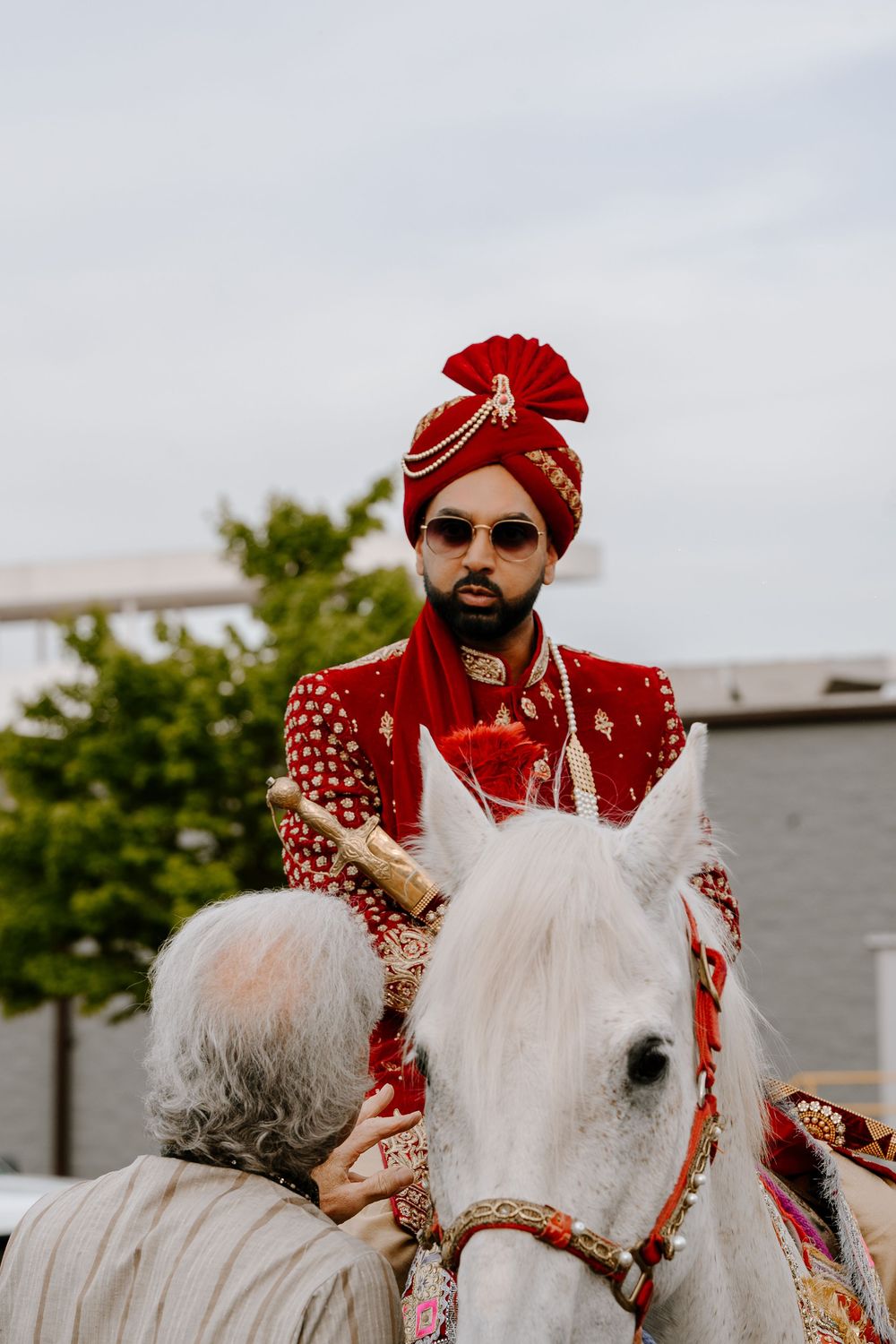 Indian groom in red sherwani and turban rides decorated white horse during traditional Baraat wedding procession.