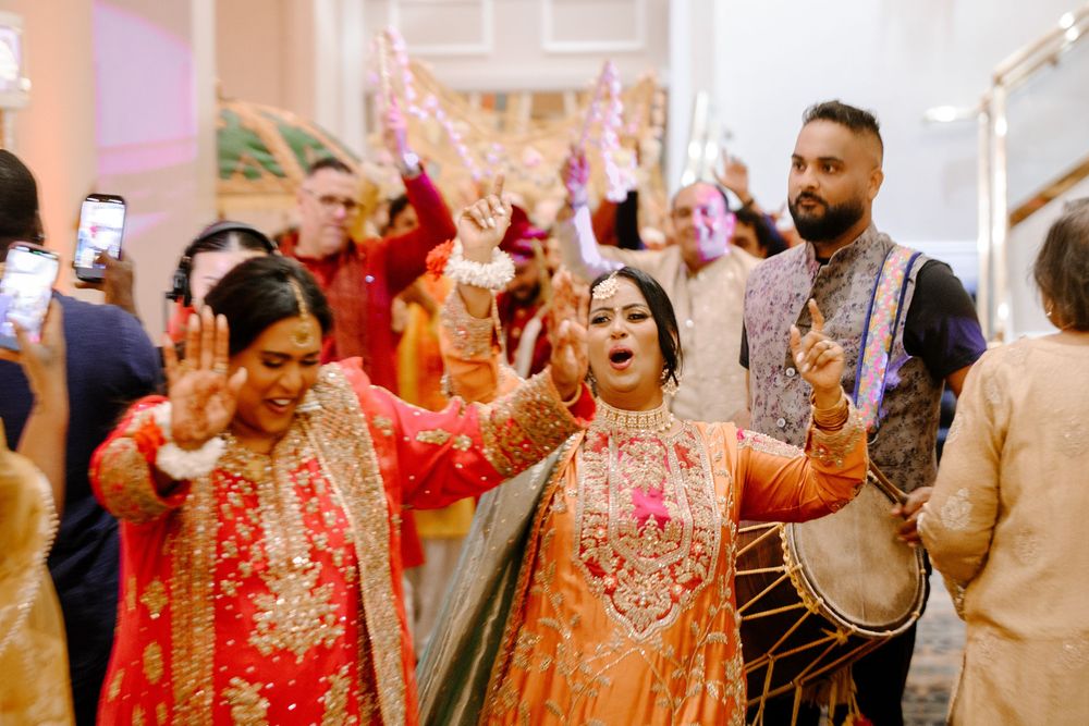 Bride in orange lehenga celebrates wedding entrance with guests dancing and drummer at vibrant South Asian ceremony.