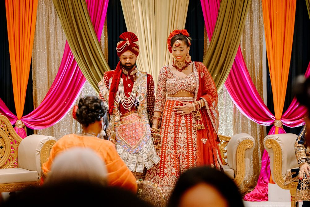 Indian bride and groom in red traditional wedding attire stand at ornate mandap with colorful pink and orange drapes.