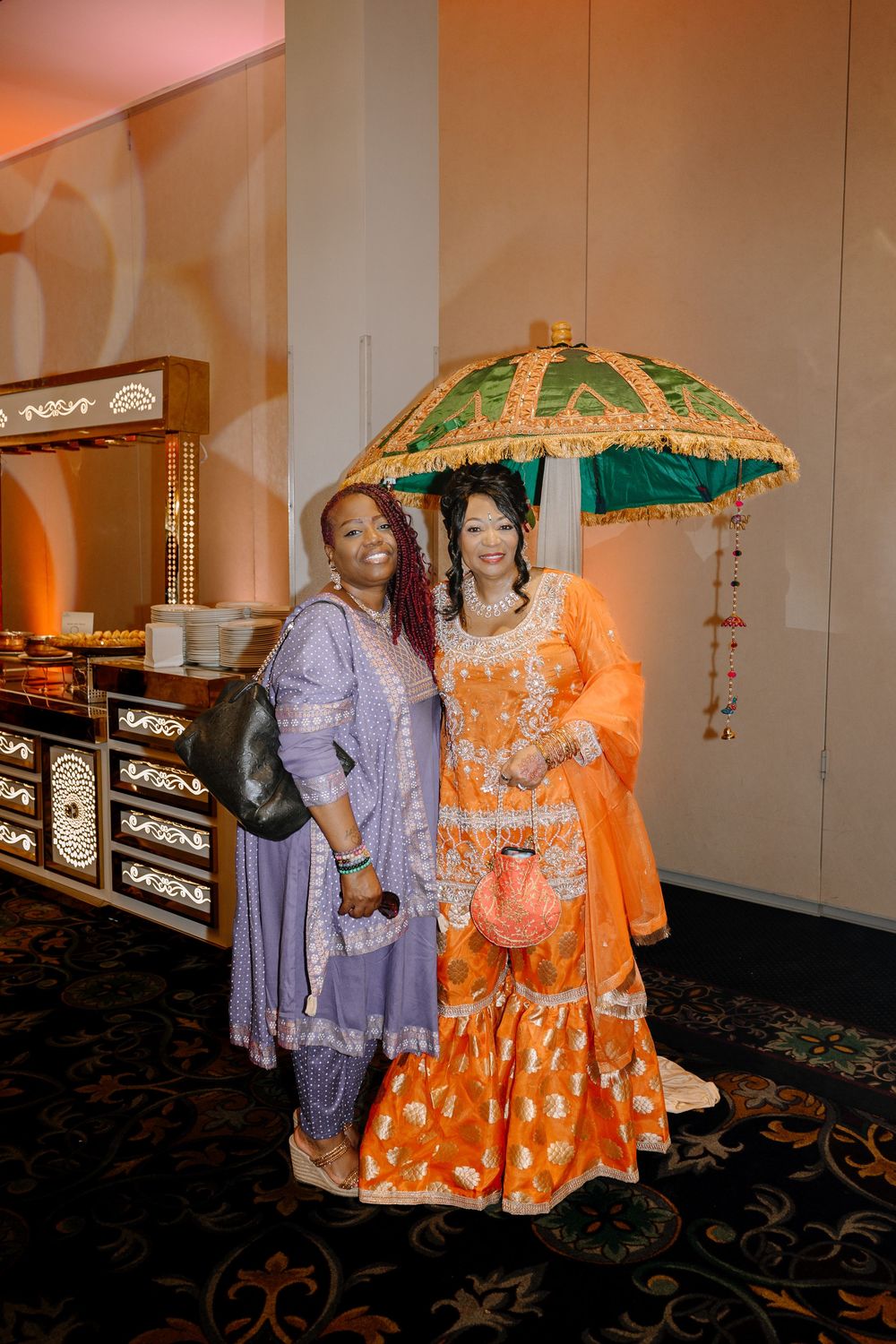 Two women in vibrant traditional South Asian attire pose together, one in orange holding a decorative green umbrella.