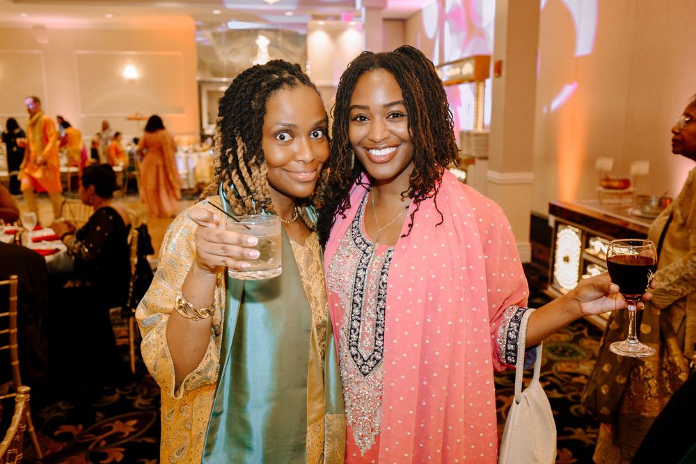 Two smiling women posing together at an elegant indoor event, wearing colorful traditional attire and holding drinks.
