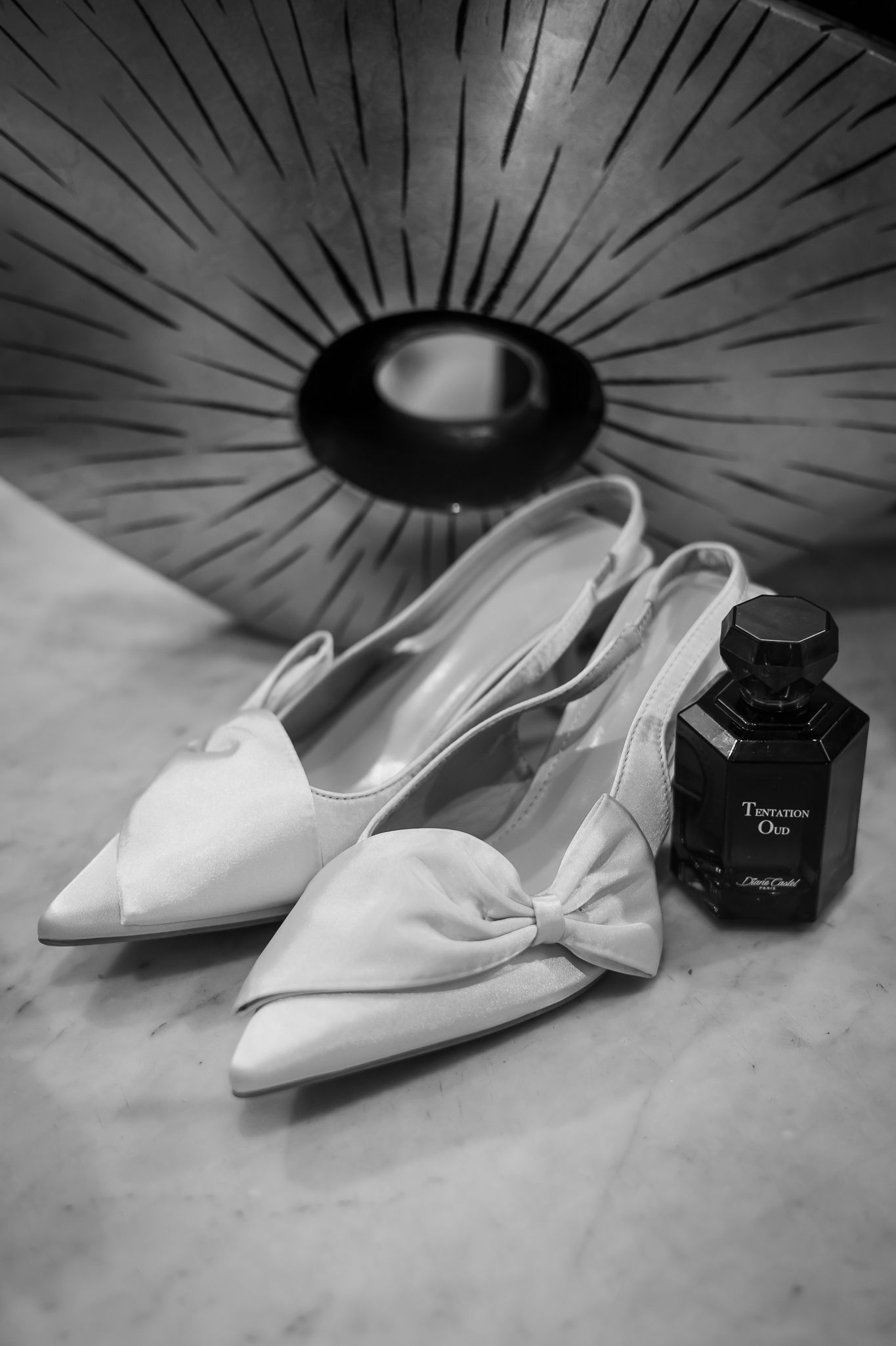 Black and white photo of white slingback bow heels beside a perfume bottle on marble surface.