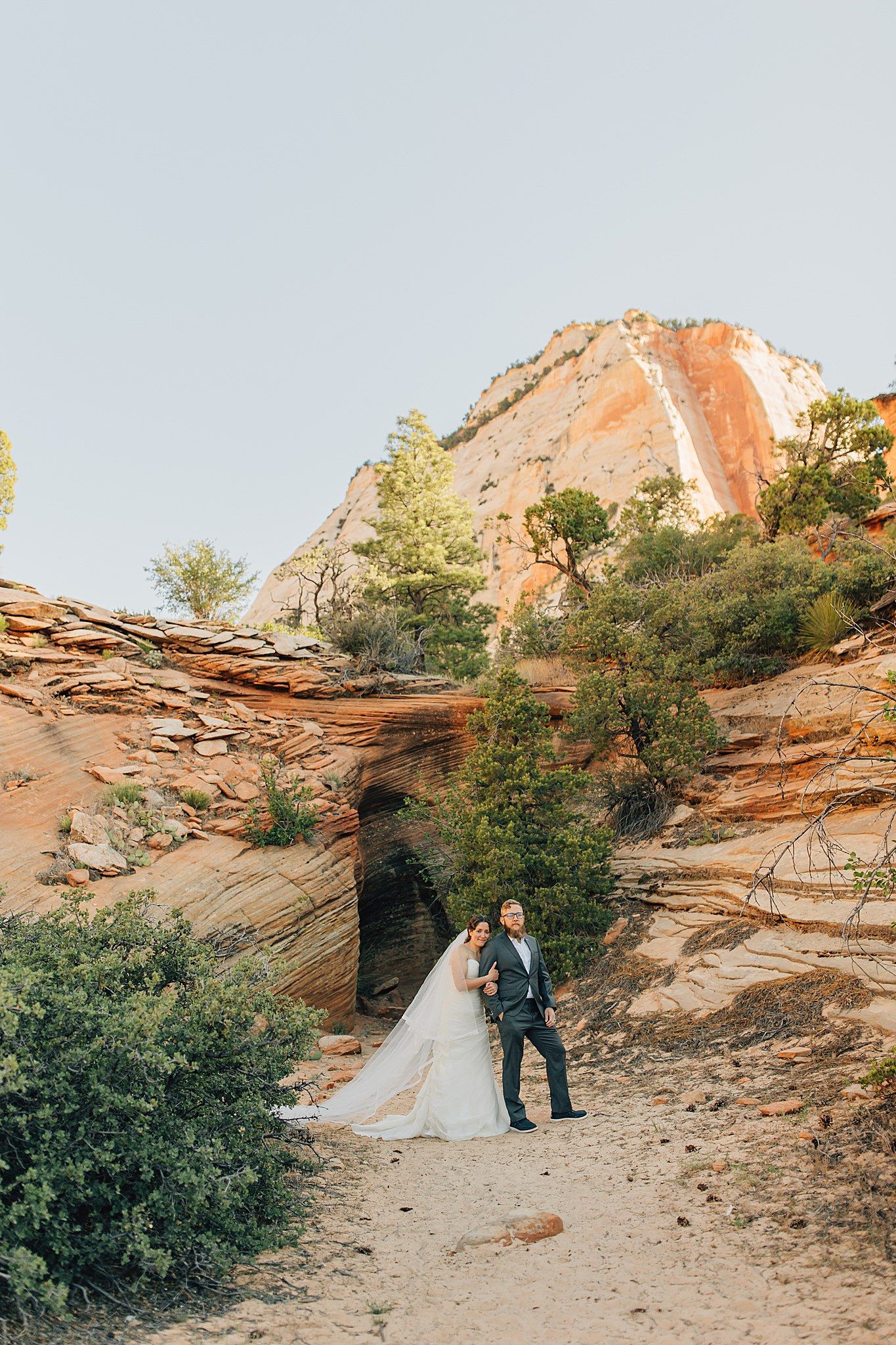 Bride and groom pose among red rock formations and green trees at Zion National Park in a stunning desert wedding photo.