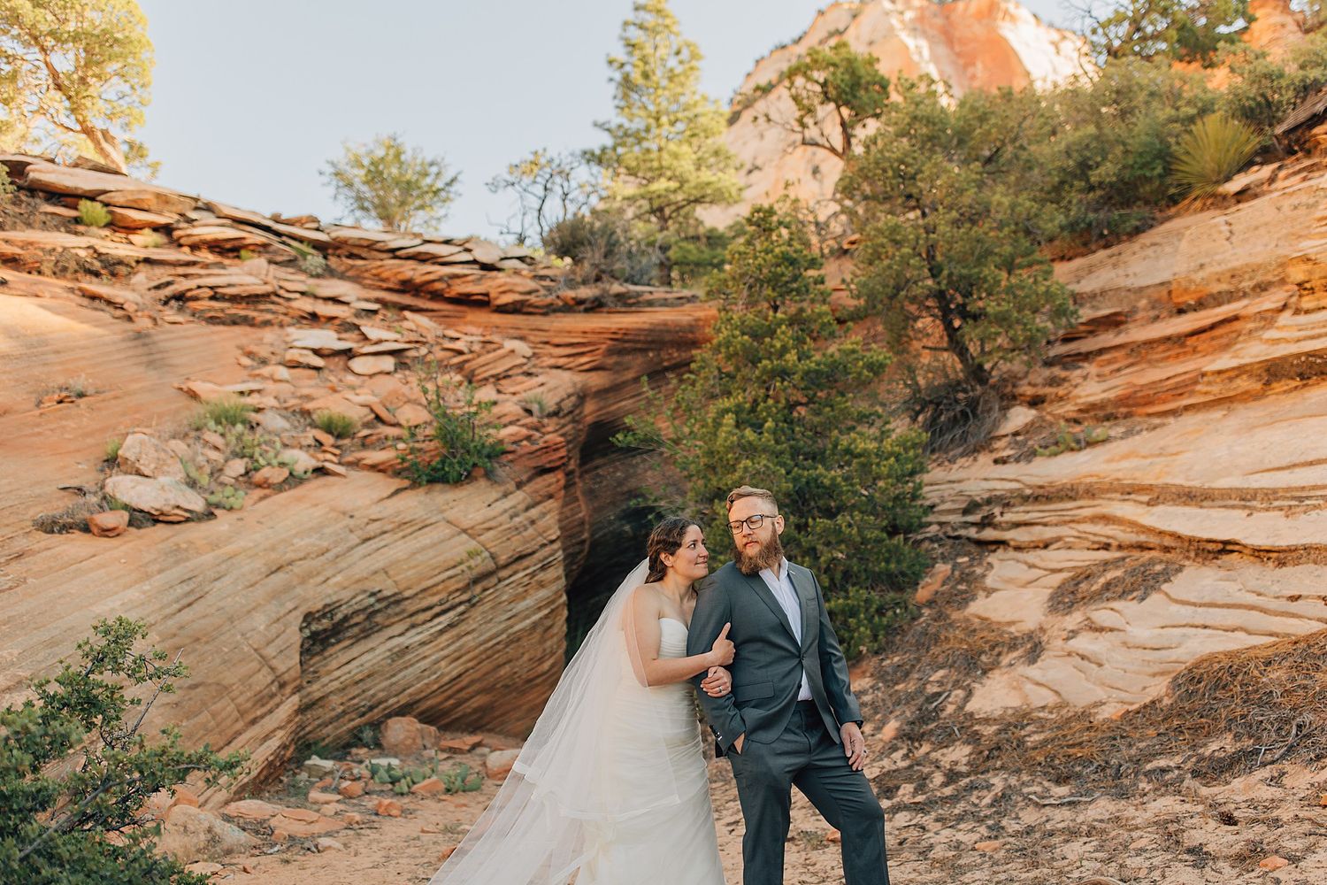 Couple in wedding attire embraces among red sandstone formations at Zion National Park at sunset.