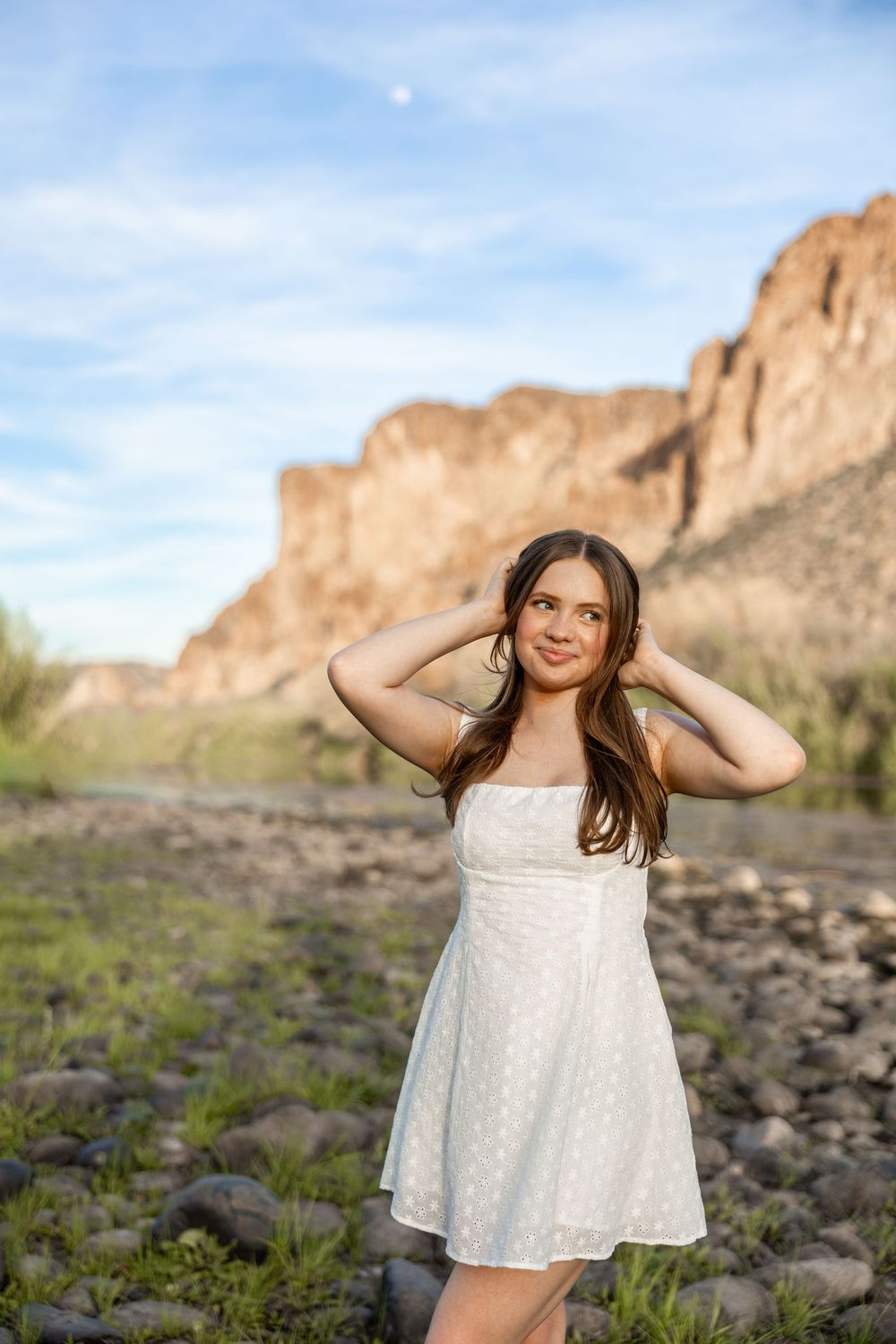 Young woman in white sundress posing with hands in hair near rocky desert canyon landscape.