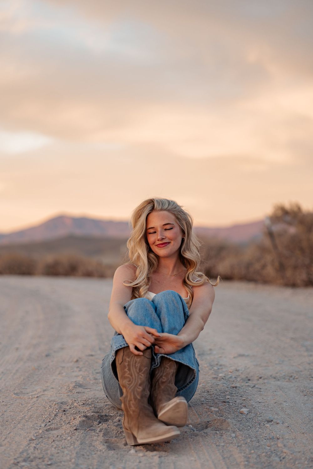 Blonde woman in jeans and cowboy boots sitting on desert road at sunset with mountains.