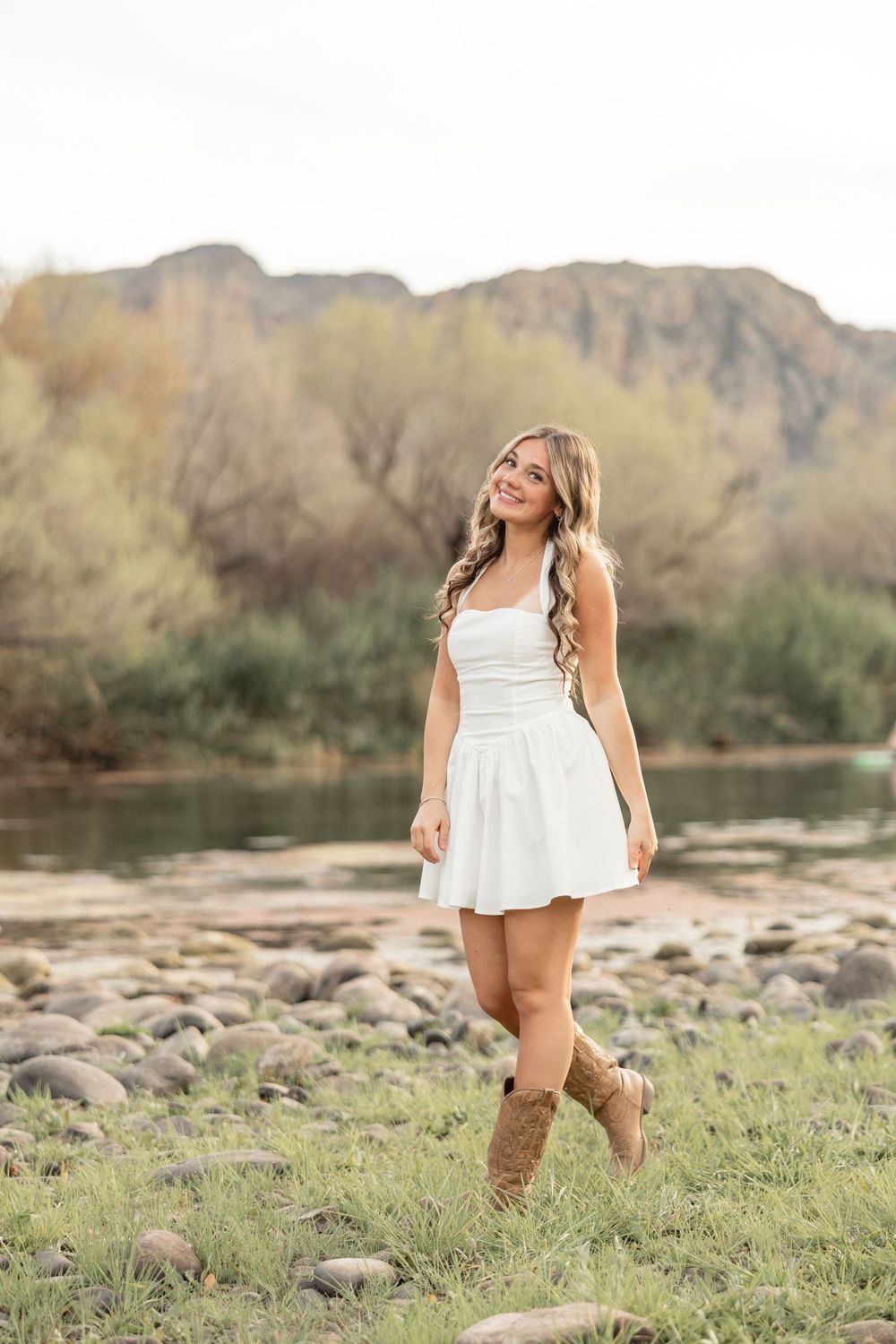 Young woman in white dress and brown cowboy boots smiling outdoors near a rocky riverbank with mountains in background.