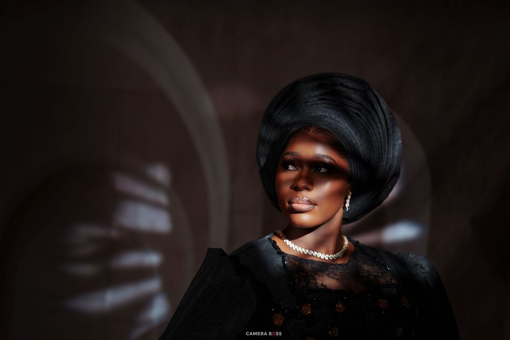 Elegant woman in black lace dress with pearl necklace and sculptural updo, posed in dramatic studio lighting.